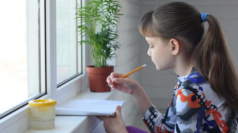Girl draws by the window with a pencil in an album