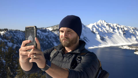 Man recording himself with snowy mountain range and lake as background