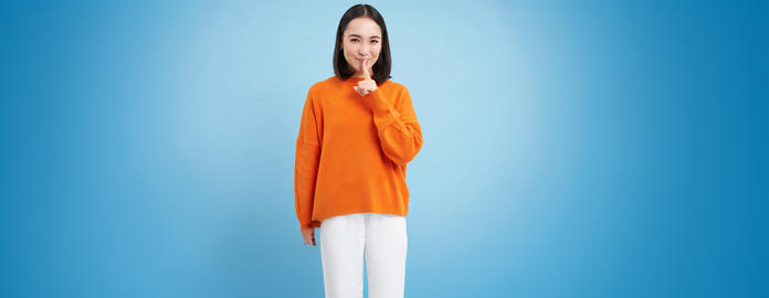 Hush its a secret. Young asian woman shushing, asks to be quiet, holds finger on lips, gossips