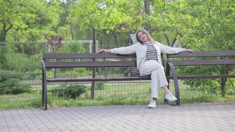 A woman in light clothing sits on a park bench with her hands behind her head