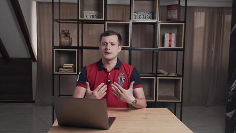 Man at table with laptop is telling something looking at camera