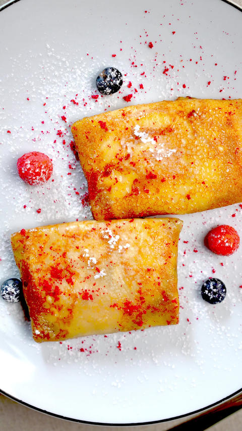 Golden nalysnyky adorned with fresh berries, powdered sugar, red crumble, sitting beside orange