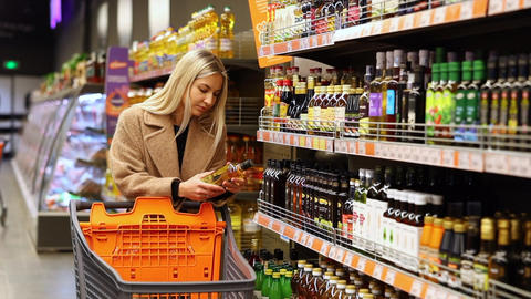 Smiling woman choosing olive oil in supermarket, holding a bottle and shopping cart