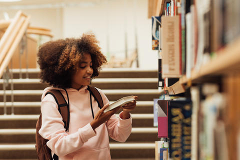Smiling girl with curly hair holding a book near bookshelf in school library