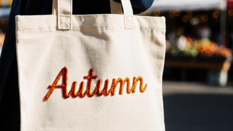 Autumn text sewn on a linen tote, outdoor market stalls in background, hdr, detailed, dynamic, intri