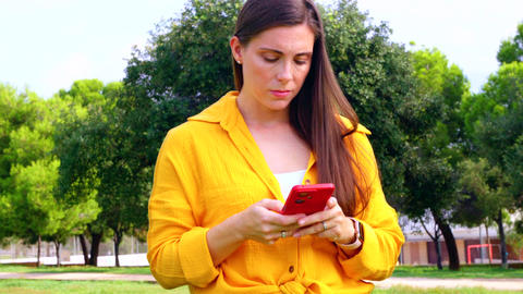 Smiling woman in yellow shirt holding smartphone and looking at camera outdoors