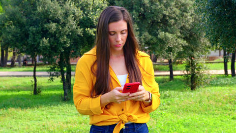 Pensive woman in yellow shirt holding smartphone outdoors thinking in park