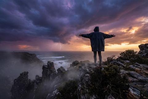Solitary man on dramatic cliff edge with outstretched arms facing lightning storm over sea