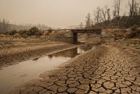 Cracked dry mud in riverbed with small stream under concrete bridge and hazy smoky sky