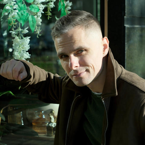 Man striking a confident pose in a sunlit urban setting with greenery in the background