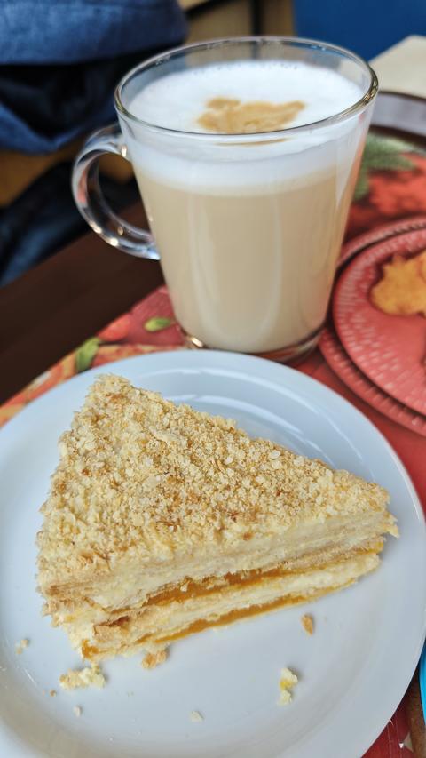 Layered cream cake slice with crumb topping served on white plate alongside glass of latte. Cozy des