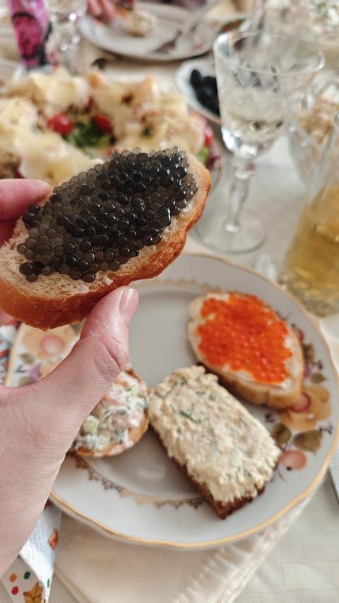 Slice of bread with black caviar held by hand over festive table with assorted appetizers and drinks
