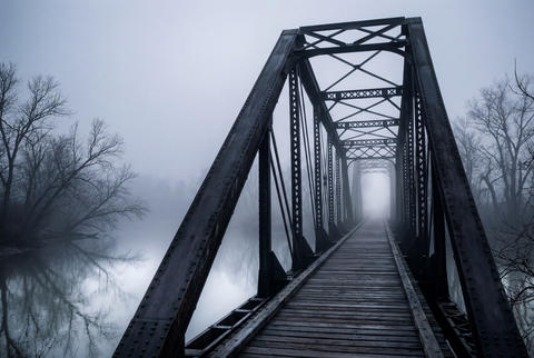 Old steel truss bridge with wooden deck crossing river into dense fog and mist