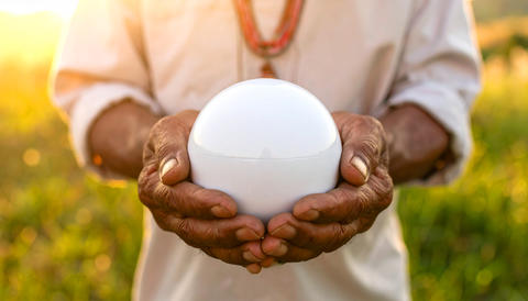 Elderly Indigenous man with trembling hands holds a sleek spherical object in a field during sunset 