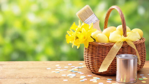 A basket full of eggs and paintbrushes with a can of paint on a wooden table with space for text