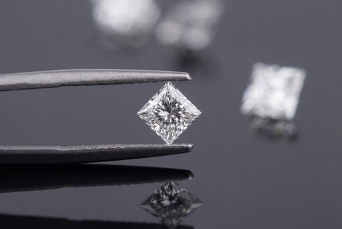Princess cut diamond held precisely by jewelry tweezers on a dark reflective surface