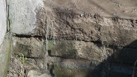 Concrete wall wastewater leak dripping from weathered curb into shadowed drain, moss and sediment