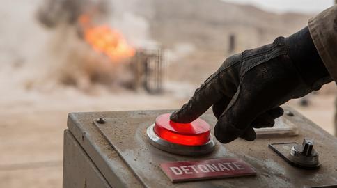 Gloved Hand Pressing Detonate Button with Explosion Background