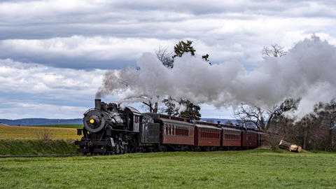 Steam Train Travels Along a Green Field Under a Cloudy Sky