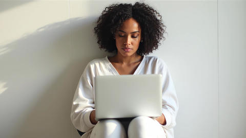 Young curly woman sitting indoors with laptop looking up smiling and blowing kiss to viewer