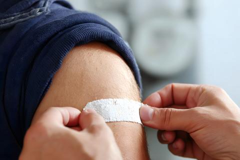 A persons hand carefully applies a small white medical bandage to the upper arm after an injection p