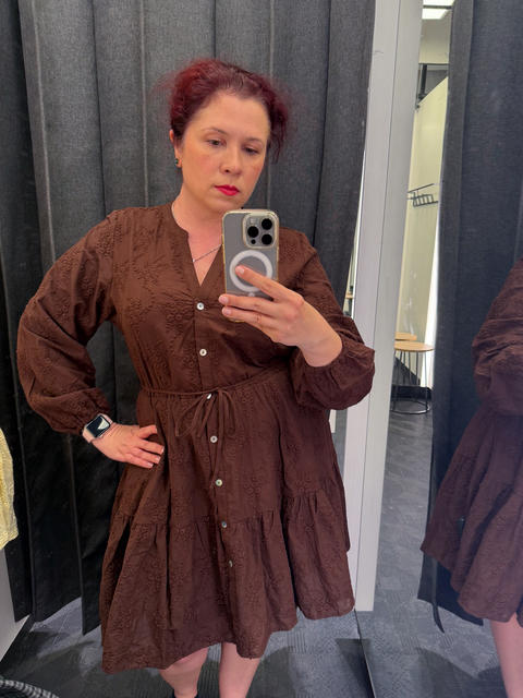 Woman takes a mirror selfie while wearing a brown embroidered dress in a fitting room. Fashion
