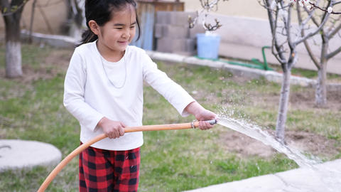 A cute Asian girl holds a hose in her hands and pours water into the garden. Funny children's games