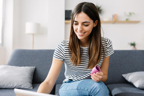 Young woman holding menstrual cup looking for use instructions on laptop