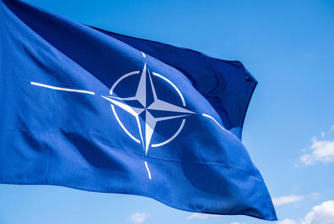 The flag of NATO waving against a clear blue sky with white clouds