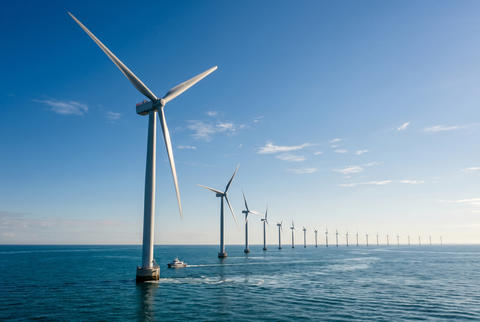 Row of offshore wind turbines in the deep blue ocean with a small boat passing by under clear sky