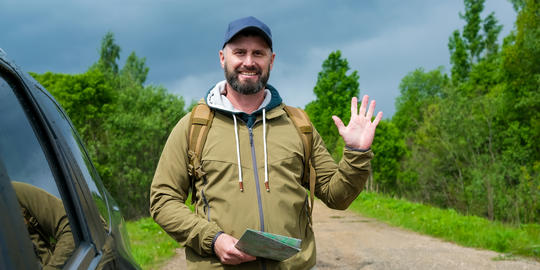 Smiling friendly bearded man with backpack stands near crossover vehicle on rural road and waves