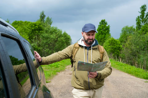 Paper map helps adult man with backpack choose route beside crossover vehicle before continuing