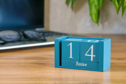 Blue desk calendar showing 14 June on wooden table with laptop glasses and plant leaf in soft