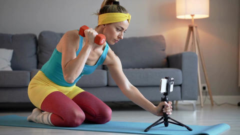 Young woman in yellow headband exercising with red dumbbell on yoga mat, medium shot