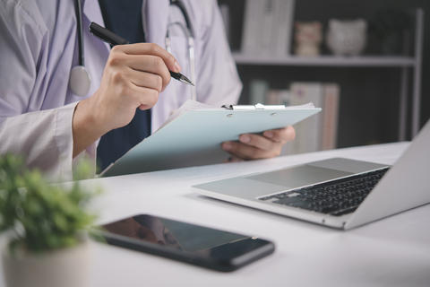 doctor writing medical record on clipboard at clinic desk with laptop and stethoscope. Physician fil