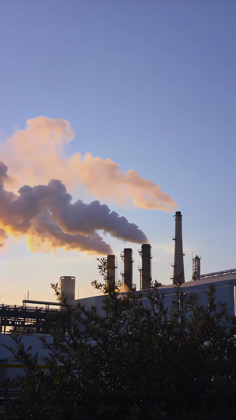 Factory smokestacks releasing dark emissions into evening sky, telephoto view of industrial plant