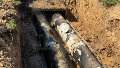 Water jet bursts from damaged heating pipe in excavated trench with muddy water and old insulation
