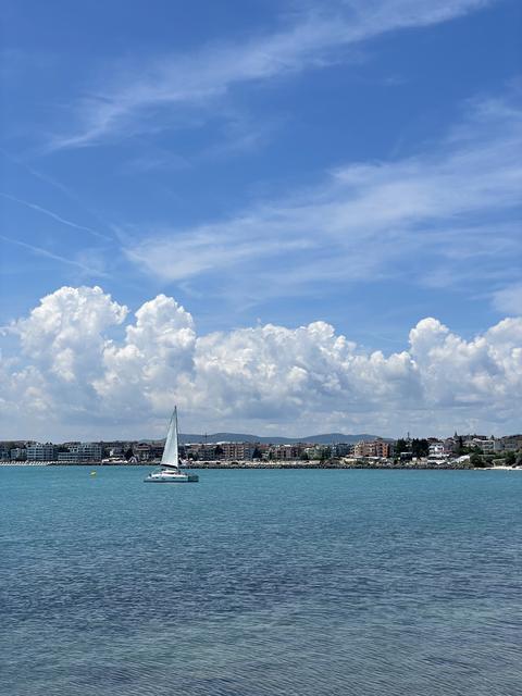 Sailboat on turquoise sea photo with coastal town, dramatic white clouds and blue sky, peaceful