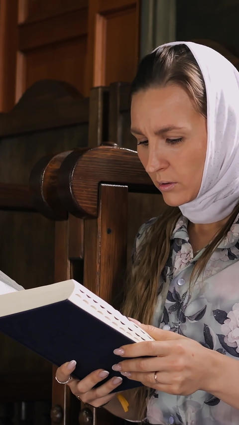 Orthodox woman reading prayer book indoors, wearing white headscarf and floral