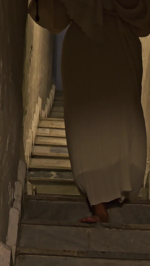 person in long beige dress walks down narrow stairway, visible only from waist