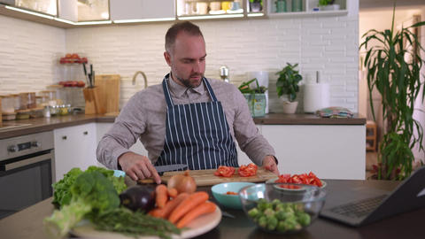 Millennial man watching online cooking tutorial and cutting tomatoes in the kitchen