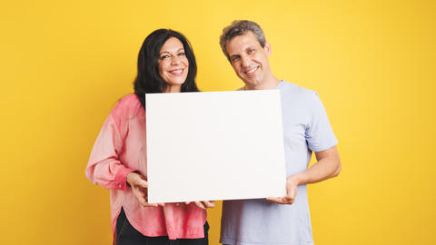 Smiling couple holding blank white board on yellow background