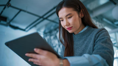 Portrait view in office interior. Asian woman wearing glasses holding tablet device. Reading