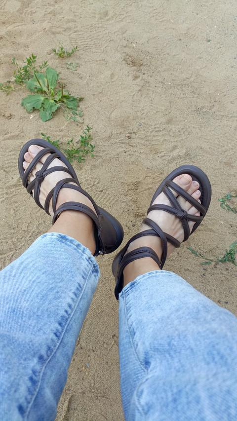 First-person top-down view of feet wearing strappy brown sandals and blue jeans, standing on sandy g