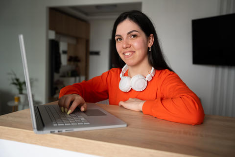 Young woman in bright room working on laptop with headphones around neck. Remote work sustainability