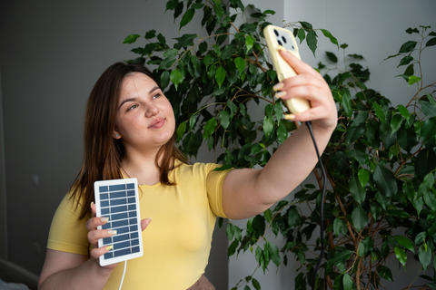 Young woman using portable solar charger to power her smartphone indoors. Renewable energy sustainab
