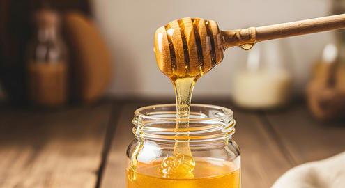 Golden honey dripping from a ladle into a glass bottle on a simple cream background