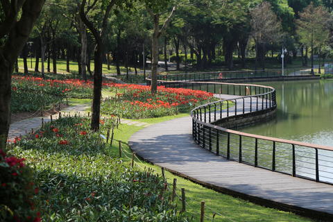 A park with red flowers and green grass, the lake is surrounded by a black metal guardrail and woode