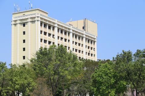 A tall building with large windows and trees in front of it, the background is a clear blue sky