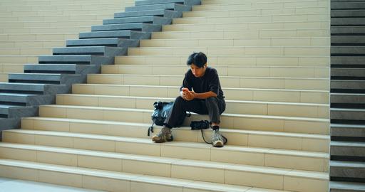 Young man sitting on broad indoor stairs, looking at smartphone beside backpack